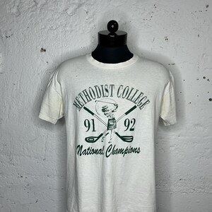 May include: White t-shirt with a green and black graphic of a woman playing golf. The graphic says "Methodist College 91 92 National Champions".