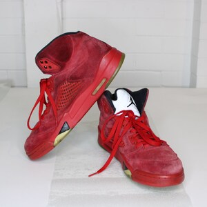 May include: A pair of red suede high-top trainers with black accents and a white logo. The trainers have a red mesh panel on the side and a white rubber sole.