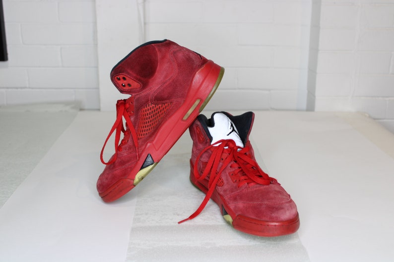 May include: A pair of red suede high-top trainers with black accents and a white logo. The trainers have a red mesh panel on the side and a white rubber sole.