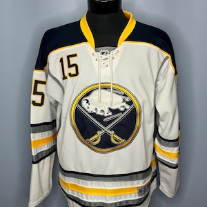 May include: White hockey jersey with navy blue and yellow accents. Features the number 15 in black on the front and sleeves. The jersey displays a team logo with a buffalo and crossed swords.