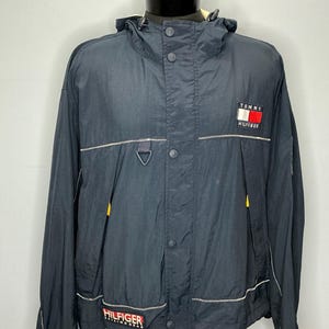 May include: Navy blue hooded jacket with the Tommy Hilfiger logo on the chest and lower front. The jacket features white stitching, yellow accents, and a button-up front. The jacket is displayed on a mannequin.