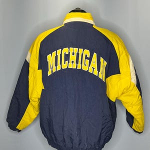 以下が含まれることがあります： 背中が黄色い文字で「MICHIGAN」と書かれた、青と黄色のビンテージのプファージャケットです。