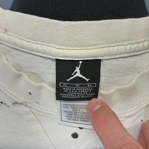 Puede incluir: Primer plano de una camiseta XL Jordan blanca con salpicaduras de pintura negra. La etiqueta de la camiseta presenta el logotipo de Jumpman y texto que indica que est&aacute; hecha en Honduras con telas de EE. UU. La etiqueta tambi&eacute;n incluye texto en franc&eacute;s y espa&ntilde;ol.