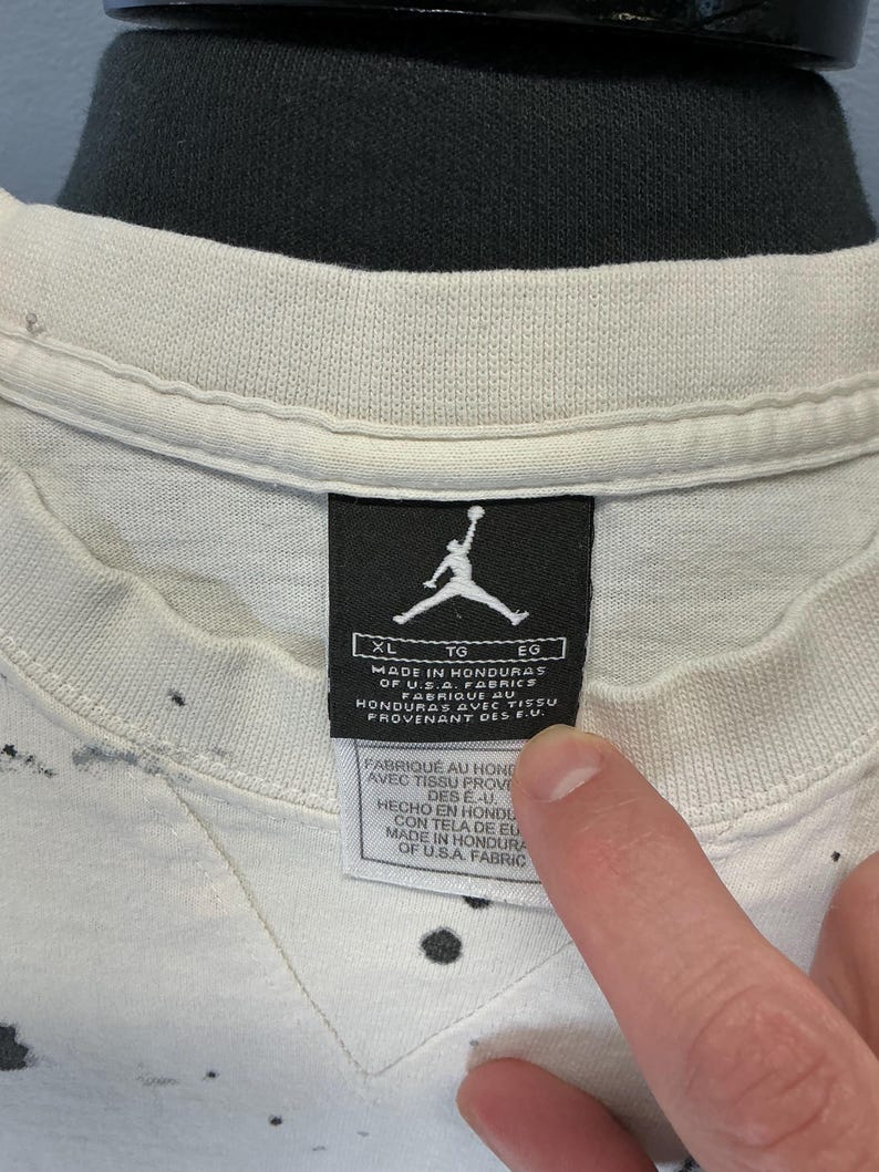 Puede incluir: Primer plano de una camiseta XL Jordan blanca con salpicaduras de pintura negra. La etiqueta de la camiseta presenta el logotipo de Jumpman y texto que indica que est&aacute; hecha en Honduras con telas de EE. UU. La etiqueta tambi&eacute;n incluye texto en franc&eacute;s y espa&ntilde;ol.