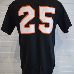 May include: Black baseball jersey with the number 25 in white with orange trim on the back. The jersey is displayed on a black mannequin. The jersey has a mesh-like texture.