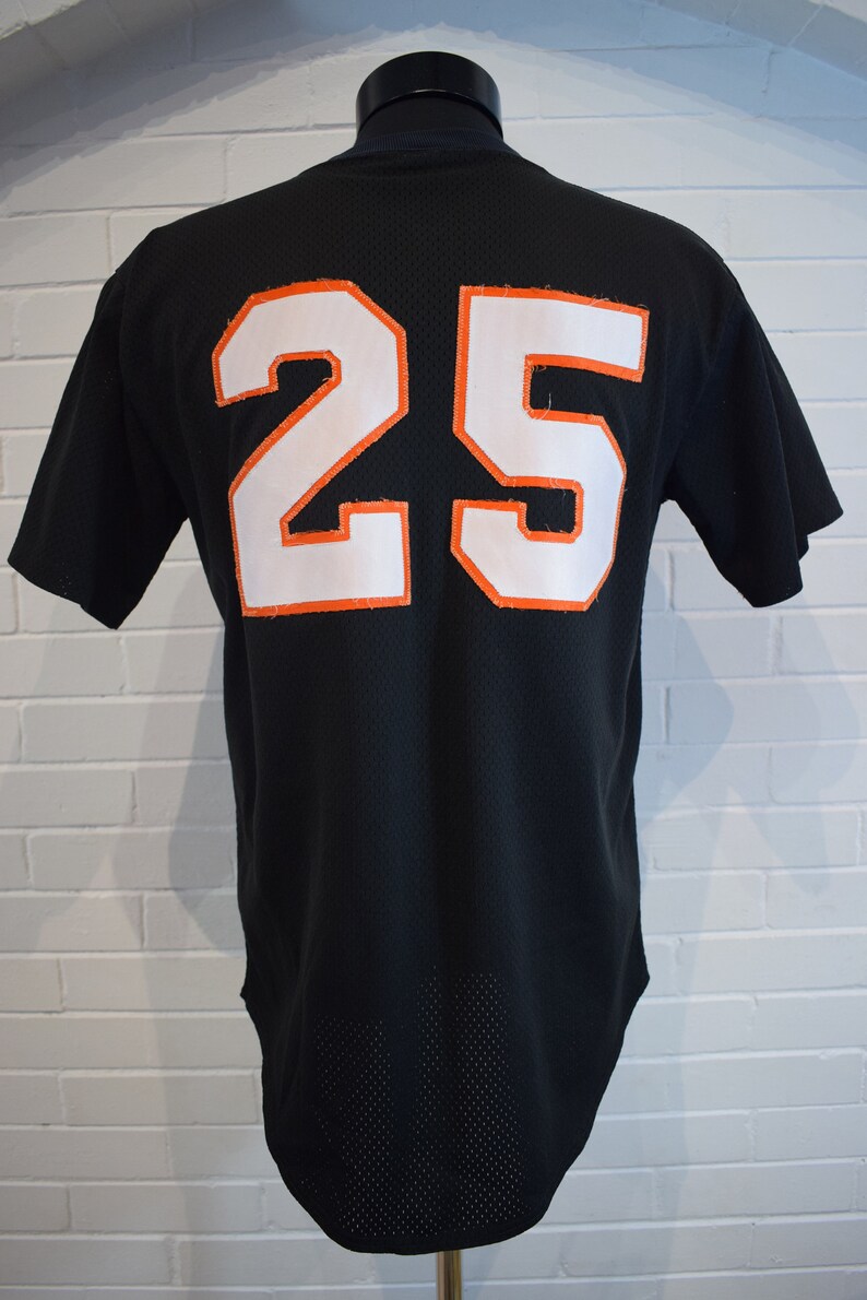 May include: Black baseball jersey with the number 25 in white with orange trim on the back. The jersey is displayed on a black mannequin. The jersey has a mesh-like texture.