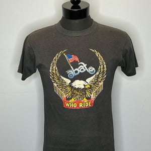 May include: A charcoal gray short-sleeve t-shirt featuring a graphic of a bald eagle with spread wings. Above the eagle is the text "abate" with a small American flag. A red banner below the eagle displays the text "WHO RIDE".