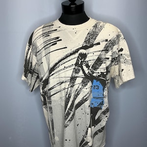Puede incluir: Camiseta color crema con un dise&ntilde;o abstracto en blanco y negro. La camiseta presenta un gr&aacute;fico de jugador de baloncesto con el n&uacute;mero 23 en azul. El dise&ntilde;o incluye salpicaduras y pinceladas negras.