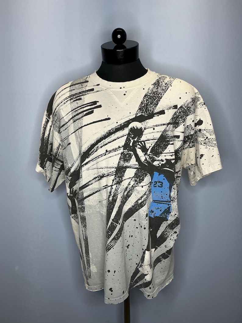 Puede incluir: Camiseta color crema con un dise&ntilde;o abstracto en blanco y negro. La camiseta presenta un gr&aacute;fico de jugador de baloncesto con el n&uacute;mero 23 en azul. El dise&ntilde;o incluye salpicaduras y pinceladas negras.