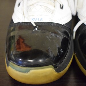 Peut inclure: Gros plan sur une paire de chaussures de basket-ball noires et blanches avec une semelle jaune. Les chaussures ont un bout en cuir verni noir et une tige blanche avec un logo bleu et blanc qui dit "XV1.5".