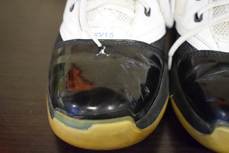 Peut inclure: Gros plan sur une paire de chaussures de basket-ball noires et blanches avec une semelle jaune. Les chaussures ont un bout en cuir verni noir et une tige blanche avec un logo bleu et blanc qui dit "XV1.5".