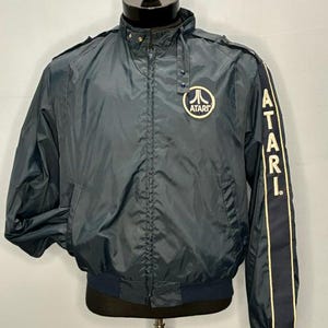 May include: A dark blue windbreaker jacket featuring a high collar with snap buttons, a full-length front zipper, and ribbed cuffs. The chest displays a circular Atari logo, while the left sleeve features the word ATARI in vertical white text.