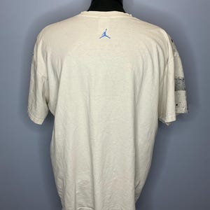 Puede incluir: Camiseta color crema con el logo Jumpman azul en la espalda. La camiseta de manga corta tiene un corte relajado. La manga derecha presenta un estampado en blanco y negro con salpicaduras de pintura. La camiseta se exhibe en un maniqu&iacute;.