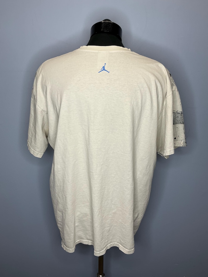 Puede incluir: Camiseta color crema con el logo Jumpman azul en la espalda. La camiseta de manga corta tiene un corte relajado. La manga derecha presenta un estampado en blanco y negro con salpicaduras de pintura. La camiseta se exhibe en un maniqu&iacute;.