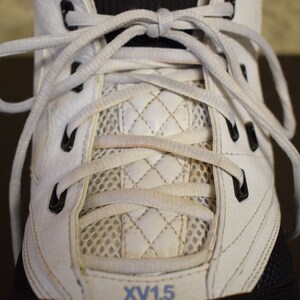 Peut inclure: Gros plan sur des baskets en cuir blanc avec des lacets blancs et des &oelig;illets noirs. Les lacets sont nou&eacute;s en croisillon. La chaussure a un panneau en mesh et un panneau matelass&eacute; blanc. Le texte "XV1.5" est visible sur la chaussure.
