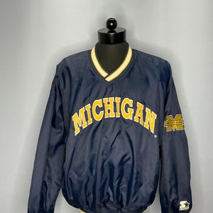 May include: A navy blue windbreaker jacket with a yellow and white striped collar and cuffs. The jacket has the word "MICHIGAN" in yellow letters on the front and a small "MICHIGAN" logo on the sleeve. The jacket is made by Starter.