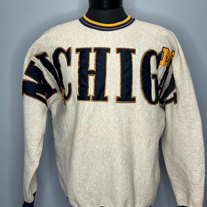 Peut inclure: Un sweat-shirt gris clair avec le mot "MICHIGAN" en lettres bleu marine et or sur la poitrine. Le sweat-shirt a des bordures bleu marine et or sur le col, les poignets et l'ourlet. Les manches ont des rayures bleu marine et or.