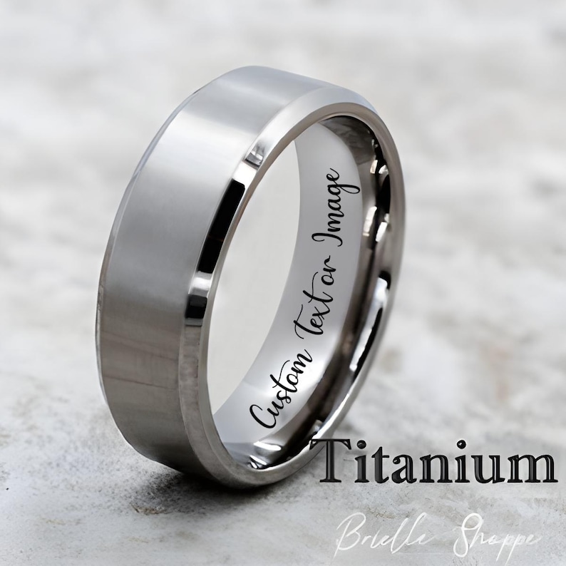 May include: A silver titanium ring with a polished finish. The ring has a flat top and beveled edges. The inside of the ring has the text "Custom Text or Image". The word "Titanium" is printed below the ring.