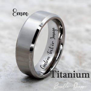 May include: A silver-colored titanium ring with a flat top and beveled edges. The ring is 8mm wide and features the text "Custom Text or Image" and "Titanium". The ring is a piece of jewelry.