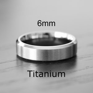 May include: A silver-colored titanium ring, 6mm wide, with a brushed finish. The ring has a flat top and beveled edges. The word "Titanium" is printed below the ring.
