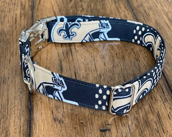 Könnte beinhalten: Ein Haustierhalsband aus Stoff mit einem schwarz-beigen Muster aus Fleur-de-Lis-Symbolen und Football-Helm-Grafiken. Das Halsband verfügt über eine silberfarbene Metallschnalle, einen D-Ring aus Metall und einen verstellbaren Metallschieber.