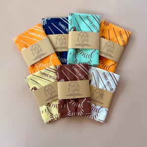 May include: A collection of colorful bandanas, each wrapped with a brown paper band. The bandanas are in various colors, including orange, navy blue, teal, yellow, brown, and white. Each bandana features a unique design and the text "THREE WOLVES TRADING POST".