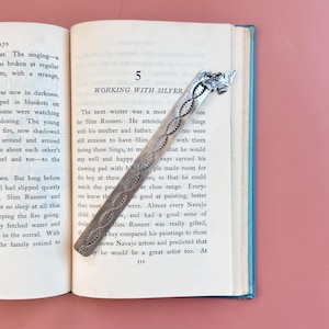 May include: A silver bookmark with an ornate design rests on an open book. The bookmark has a decorative pattern and a detailed top. The book is open to a page with text and the number 5.