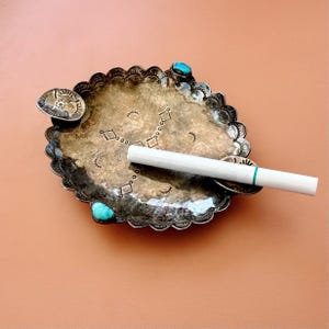 May include: A silver-coloured ashtray with a scalloped edge, decorated with turquoise stones and stamped geometric designs. A lit cigarette rests on the ashtray. The ashtray has a small, round rest for a cigarette. The background is a solid, muted orange.