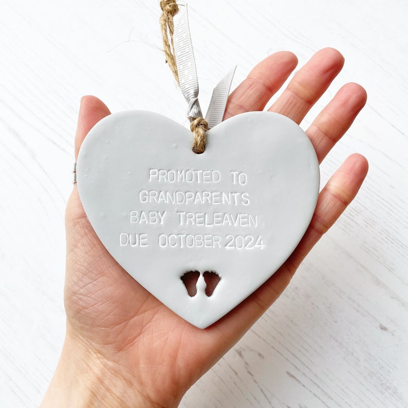 Grey clay hanging heart with baby feet cut out of the bottom and white personalisation, the heart is personalised with PROMOTED TO GRANDPARENTS BABY TRELEAVEN DUE OCTOBER 2024