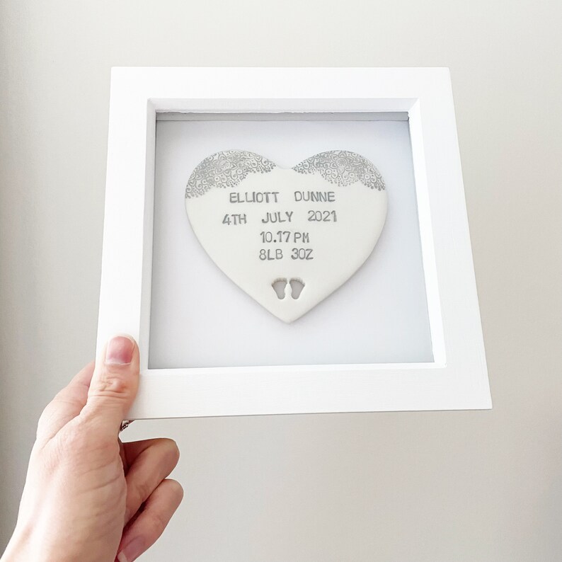 Personalised baby keepsake gift, pearlised white clay heart with lace edge top and baby feet cut out at the bottom of the heart in a white box frame, the heart is personalised with the baby’s name, date of birth, weight and time