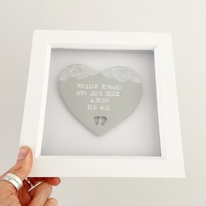 Personalised baby keepsake gift, grey clay heart with baby feet cut out at the bottom of the heart and white lace edge detail to the top in a white box frame, the heart is personalised with the baby’s name, date of birth, weight and time