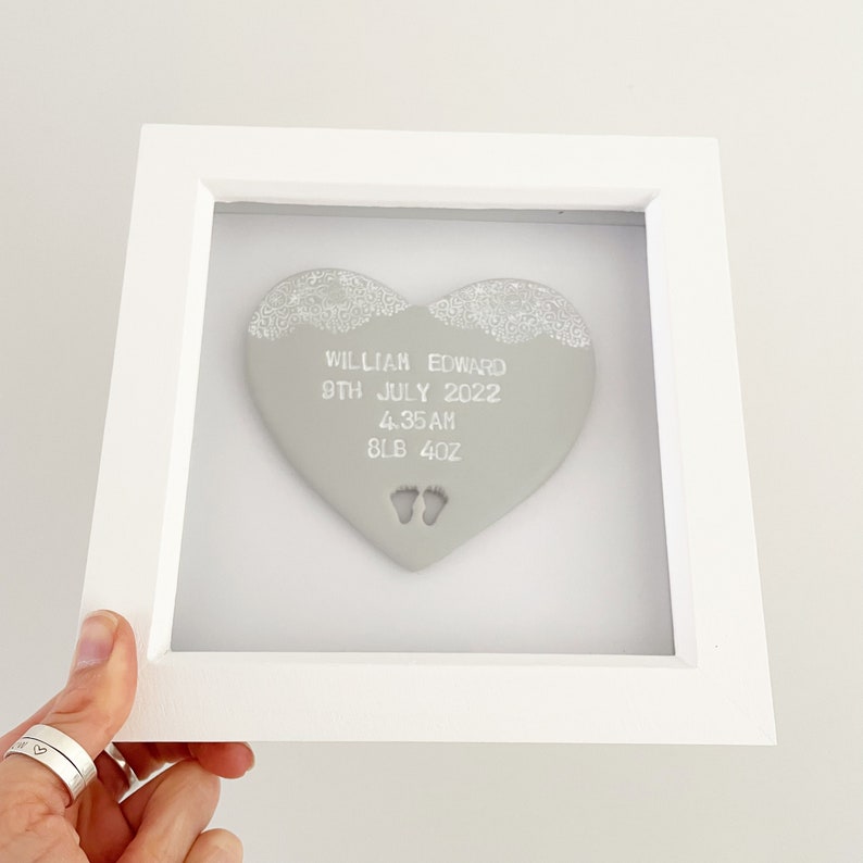 Personalised baby keepsake gift, grey clay heart with baby feet cut out at the bottom of the heart and white lace edge detail to the top in a white box frame, the heart is personalised with the baby’s name, date of birth, weight and time