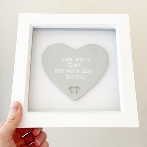 Personalised baby keepsake gift, grey clay heart with baby feet cut out at the bottom of the heart in a white box frame, the heart is personalised with the baby’s name, date of birth, weight and time