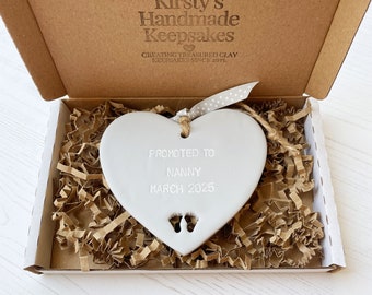 May include: White ceramic heart-shaped ornament with the text "Promoted to Nanny March 2025" and a pair of tiny footprints.