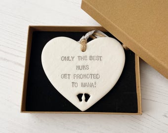 May include: White ceramic heart-shaped ornament with the text "Only the best mums get promoted to Nana!" and two small footprints.