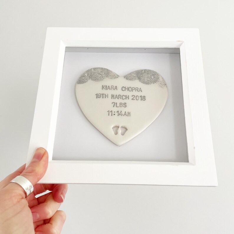 Personalised baby keepsake gift, pearlised white clay heart with lace edge top and baby feet cut out at the bottom of the heart in a white box frame, the heart is personalised with the baby’s name, date of birth, weight and time