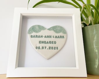 May include: A framed heart-shaped ceramic plaque with the words "SARAH-ANN & MARK ENGAGED 06.07.2025" in a soft green font. The heart has a decorative lace pattern at the top. The frame is white.