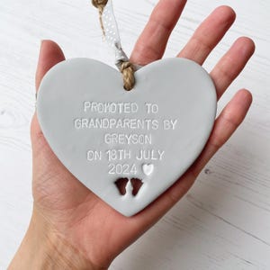 Grey clay hanging heart with baby feet cut out of the bottom and white personalisation, the heart is personalised with PROMOTED TO GRANDPARENTS BY GREYSON ON  18TH JULY 2024