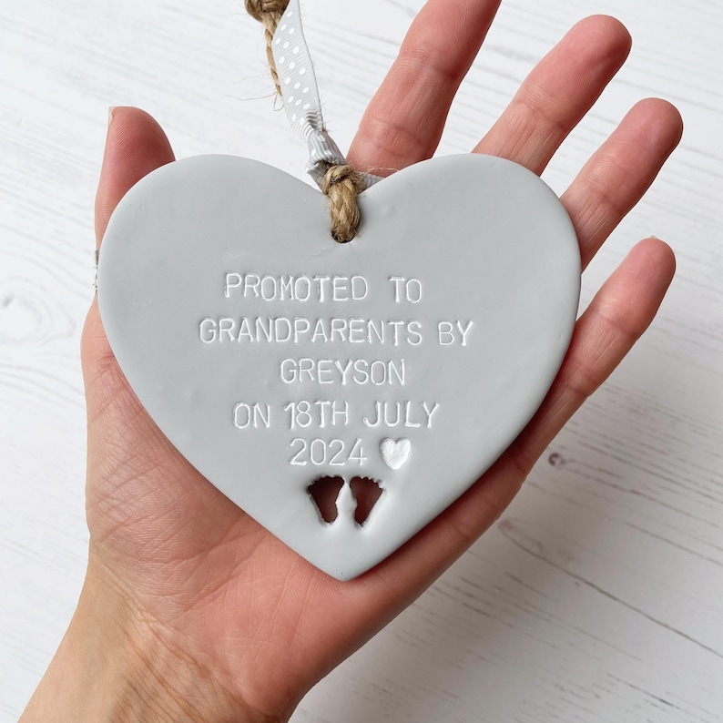 Grey clay hanging heart with baby feet cut out of the bottom and white personalisation, the heart is personalised with PROMOTED TO GRANDPARENTS BY GREYSON ON  18TH JULY 2024