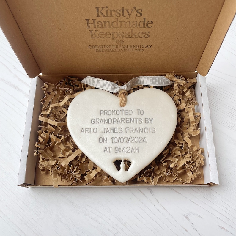 Pearlised white clay hanging heart with baby feet cut out of the bottom and grey personalisation, the heart is personalised with PROMOTED TO GRANDPARENTS BY ARLO JAMES FRANCIS ON 10/07/2024 AT 9:42 AM