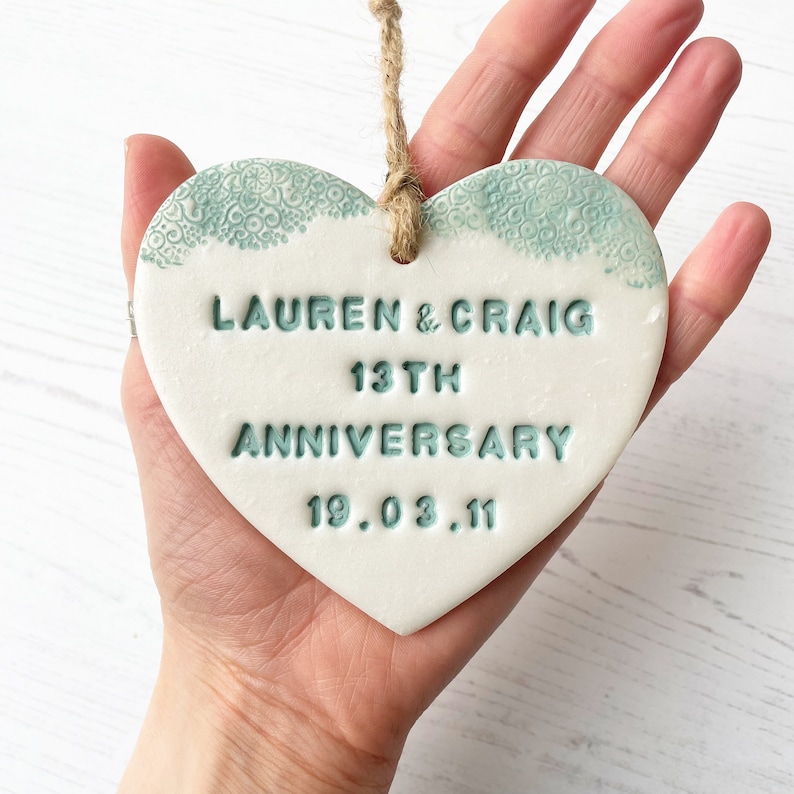 Personalised 13th anniversary gift, pearlised white clay hanging heart with a sage green lace edge at the top of the heart, the heart is personalised with LAUREN & CRAIG 13TH ANNIVERSARY 19.03.11