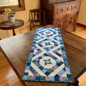 May include: A quilted table runner with a geometric pattern in shades of purple, green, and white. The runner features a series of diamond shapes with a star in the center of each diamond.