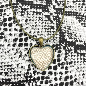 May include: A heart-shaped pendant necklace with a beige and white patterned design inside the heart. The pendant is made of metal and has a brown cord.