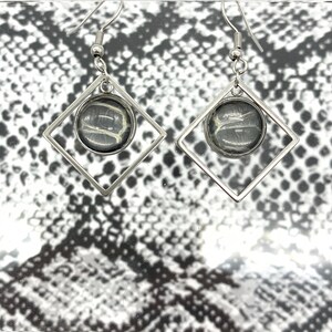 May include: A pair of silver earrings with a square frame and a round, clear glass pendant featuring a snakeskin pattern.