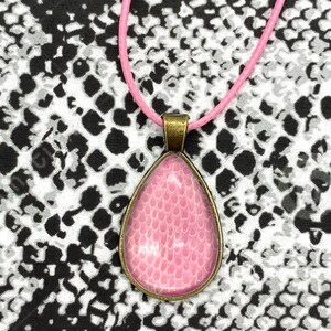 Peut inclure: Un collier avec un pendentif en forme de larme rose à surface texturée, suspendu à un cordon rose sur un fond à motifs noir et blanc.