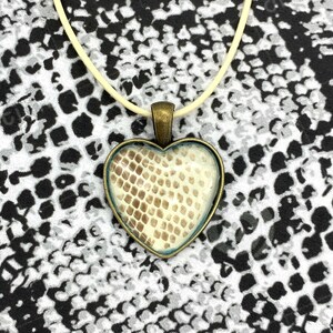 Peut inclure: Un collier avec un pendentif en forme de cœur contenant un motif de peau de serpent à l'intérieur du verre. Le pendentif est en bronze et est suspendu à un cordon blanc.