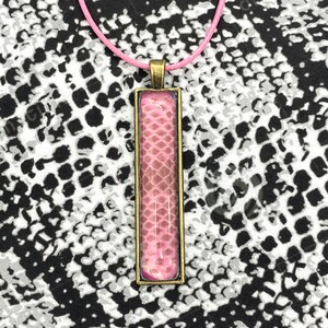 Peut inclure: Un collier avec un pendentif en peau de serpent rose dans un cadre bronze. Le collier est sur un fond à motifs blanc et noir.