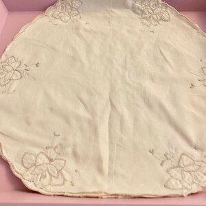 May include: A white, round tablecloth with a scalloped edge and delicate floral embroidery. The embroidery is in a light grey colour and features a repeating pattern of flowers and leaves.