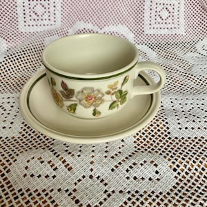 May include: A cream-coloured teacup and saucer set with a floral pattern. The cup has a green trim and a handle. The saucer is round and matches the cup's colour and design. The floral pattern includes white and yellow flowers with green leaves.