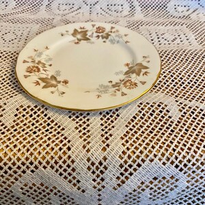 May include: A small, cream-coloured plate with a gold rim. It features a floral design in brown and tan hues. This vintage plate is likely antique.